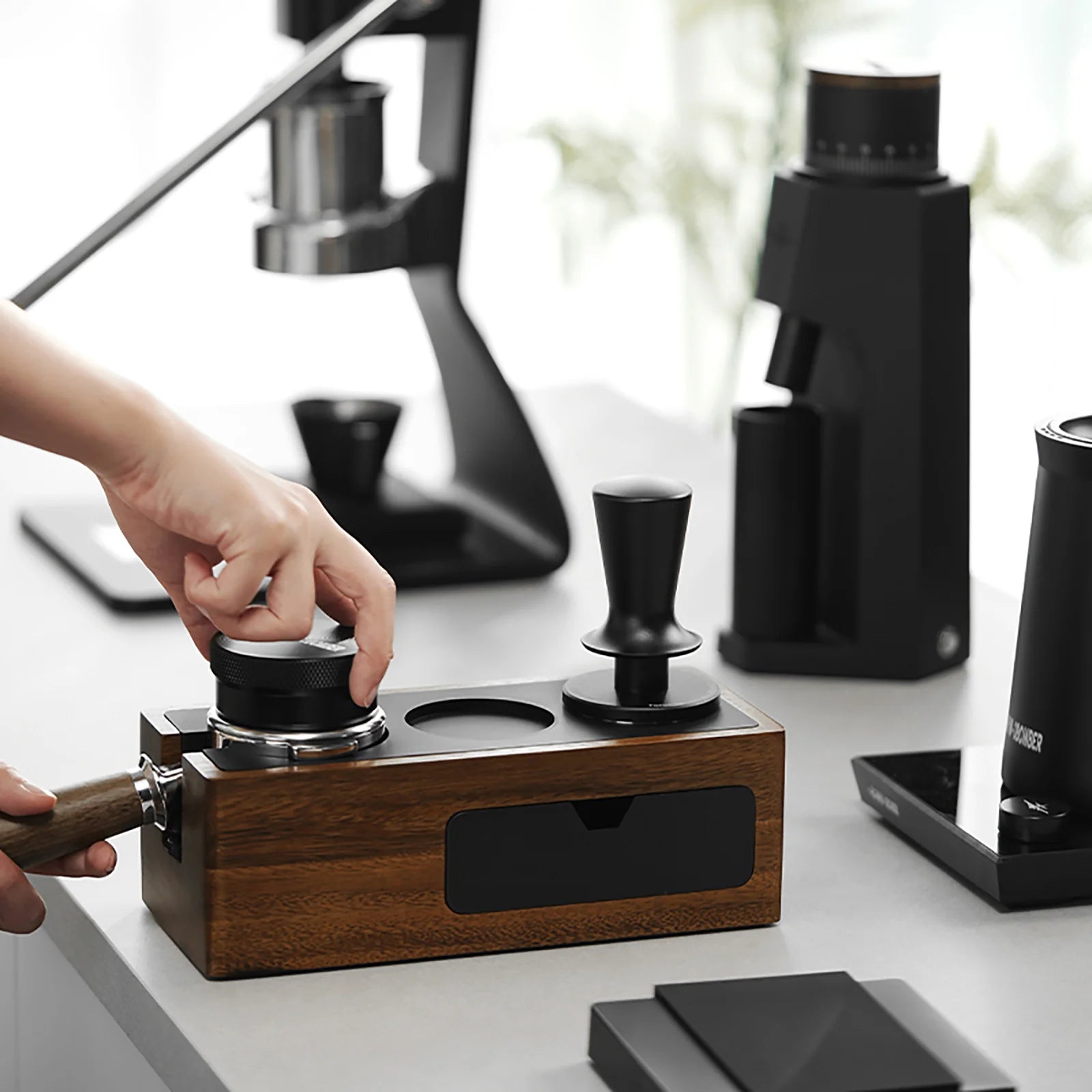 MHW-3BOMBER walnut tamper station with tamper and distributor on a counter, espresso machine in the background.