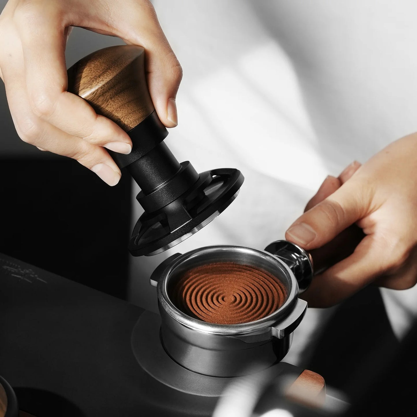 Using the MHW-3BOMBER Armor Series Coffee Tamper 2.0, a user applies constant pressure to press finely ground coffee with precision into a portafilter, creating a spiral pattern while focusing on the essential tools and techniques for espresso preparation.