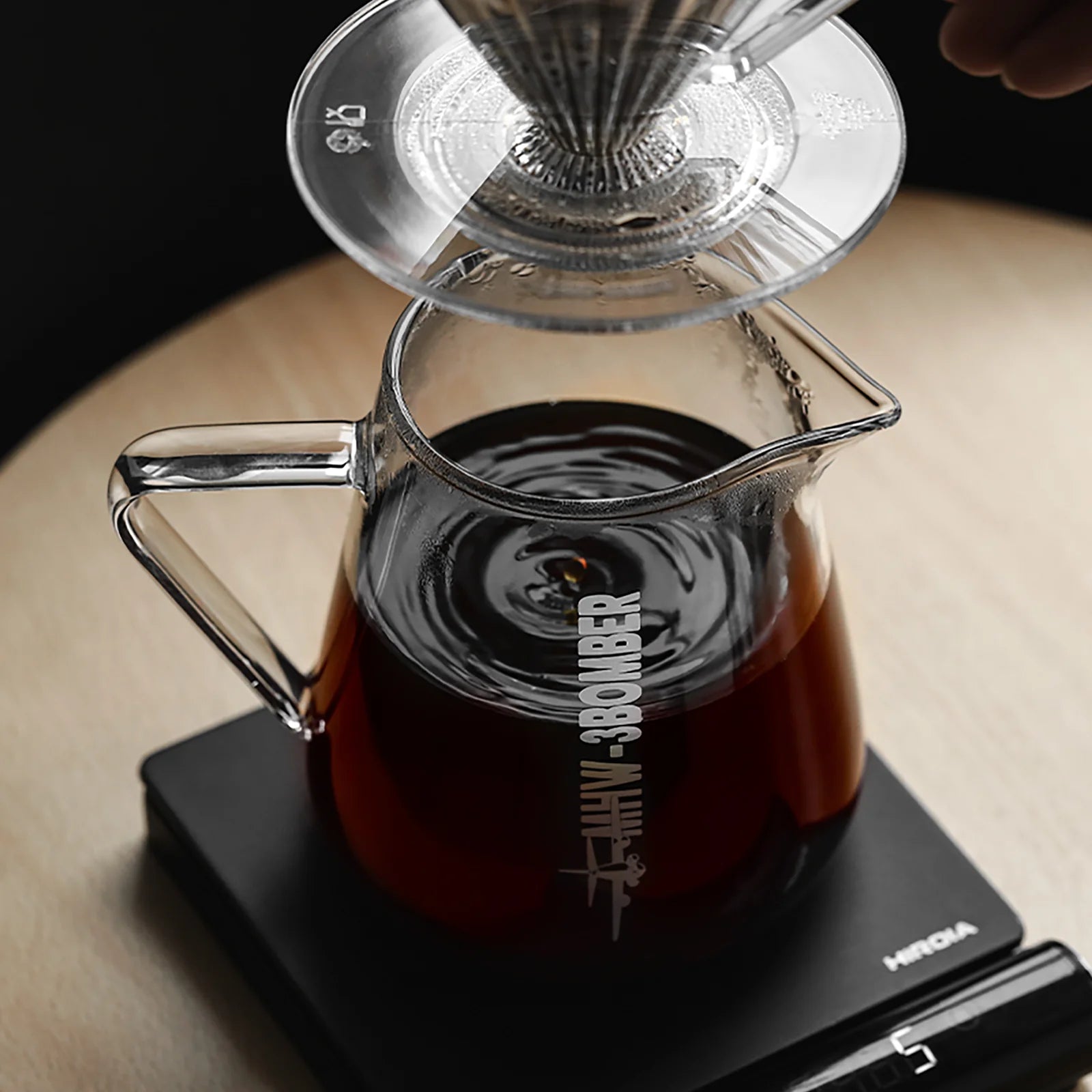 A close-up of the MHW-3BOMBER Elf Coffee Server, crafted from borosilicate glass, filled with dark coffee on a scale. A clear dripper and metallic filter sit atop, allowing coffee to drip seamlessly into the carafe against a light wooden background.