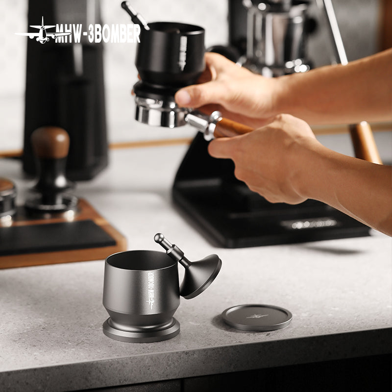 In a sleek industrial kitchen, a person skillfully uses a coffee tamper on a portafilter, surrounded by appliances like a scale and grinder. They expertly use the MHW-3BOMBER Espresso Coffee Powder Dosing Funnel, echoing the precision of professional baristas.