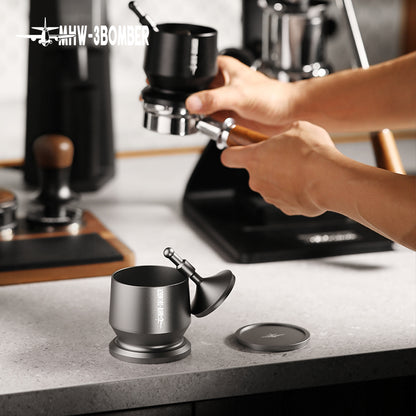 In a sleek industrial kitchen, a person skillfully uses a coffee tamper on a portafilter, surrounded by appliances like a scale and grinder. They expertly use the MHW-3BOMBER Espresso Coffee Powder Dosing Funnel, echoing the precision of professional baristas.