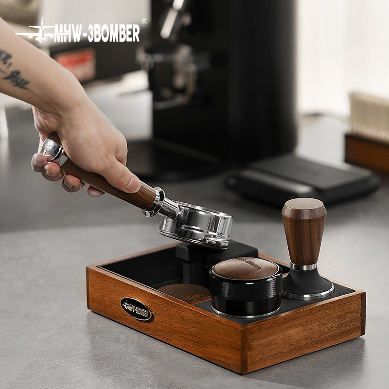 MHW-3BOMBER espresso tamping station with portafilter, tamper, and knock box. Wood and black finish on counter.