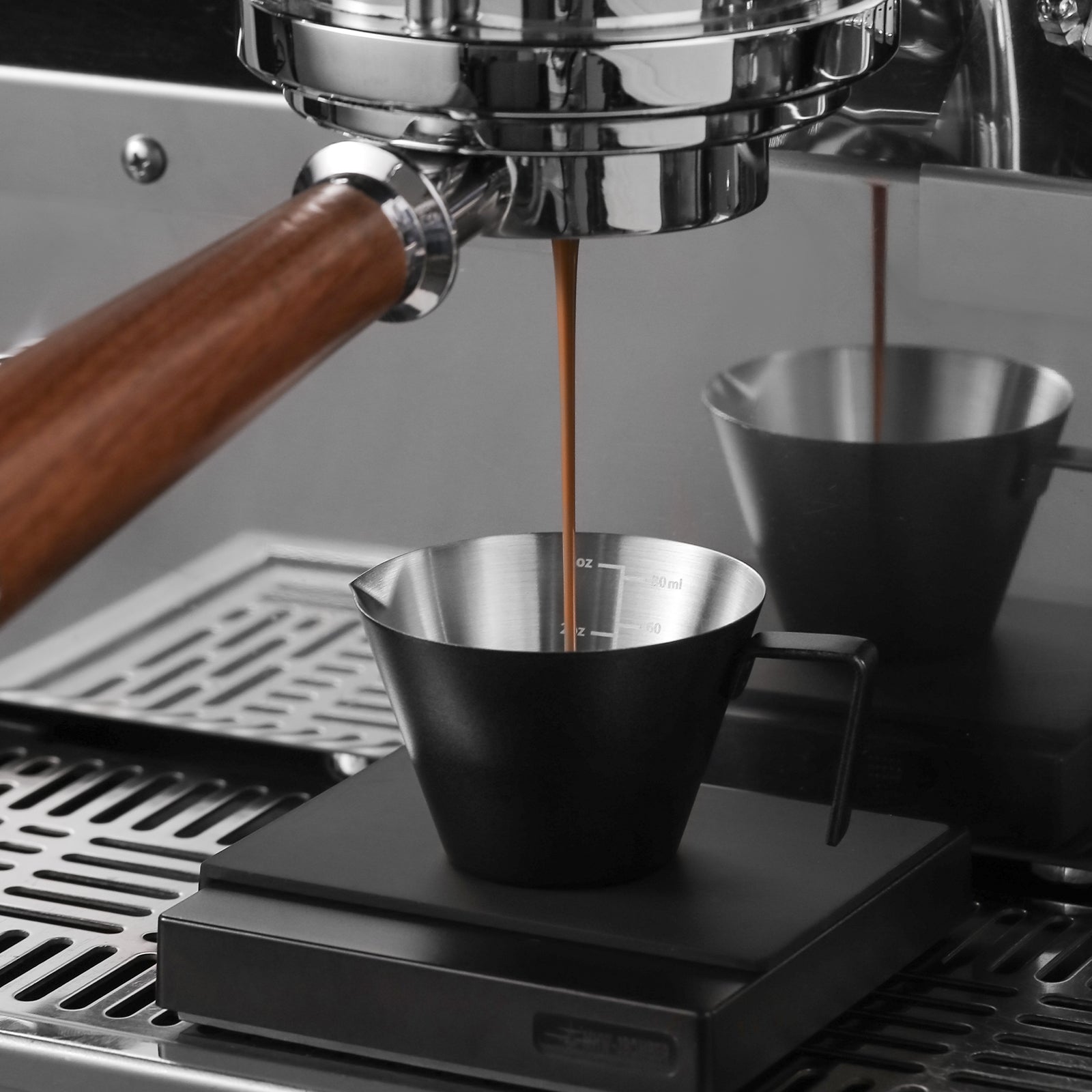 The MHW-3BOMBER espresso machine elegantly pours coffee into its sleek Stainless Steel Measuring Cup on an electronic scale. With a wooden handle and metallic parts, it shines brightly against the reflective surface, showcasing precision in coffee brewing.