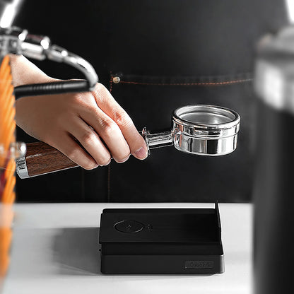 A person holds a portafilter beside the MHW-3BOMBER Espresso Scale Cube Coffee Scale 2.0 on the counter. The blurred background shows a coffee machine and someone in an apron, suggesting the scales USB rechargeability is useful for busy barista days.