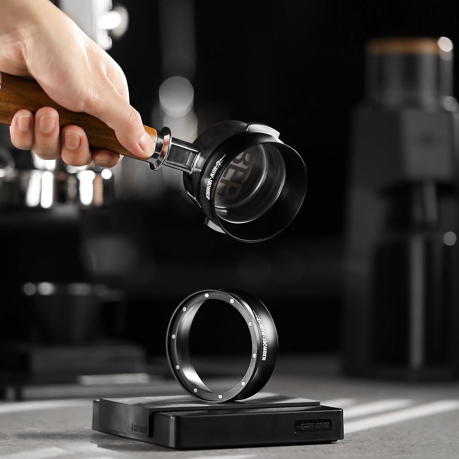 A hand holds a portafilter with a wooden handle and an MHW-3BOMBER Espresso Magnetic Coffee Dosing Funnel over a metal ring on a black platform, likely part of an espresso machine setup, with blurred coffee-making equipment in the background.