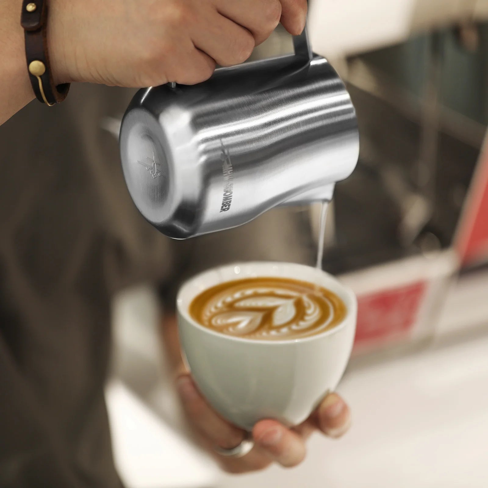 On a white countertop, someone elegantly pours steamed milk from an MHW-3BOMBER Turbo Milk Pitcher Glossy into a coffee cup, skillfully crafting leaf-shaped latte art while wearing a brown bracelet.