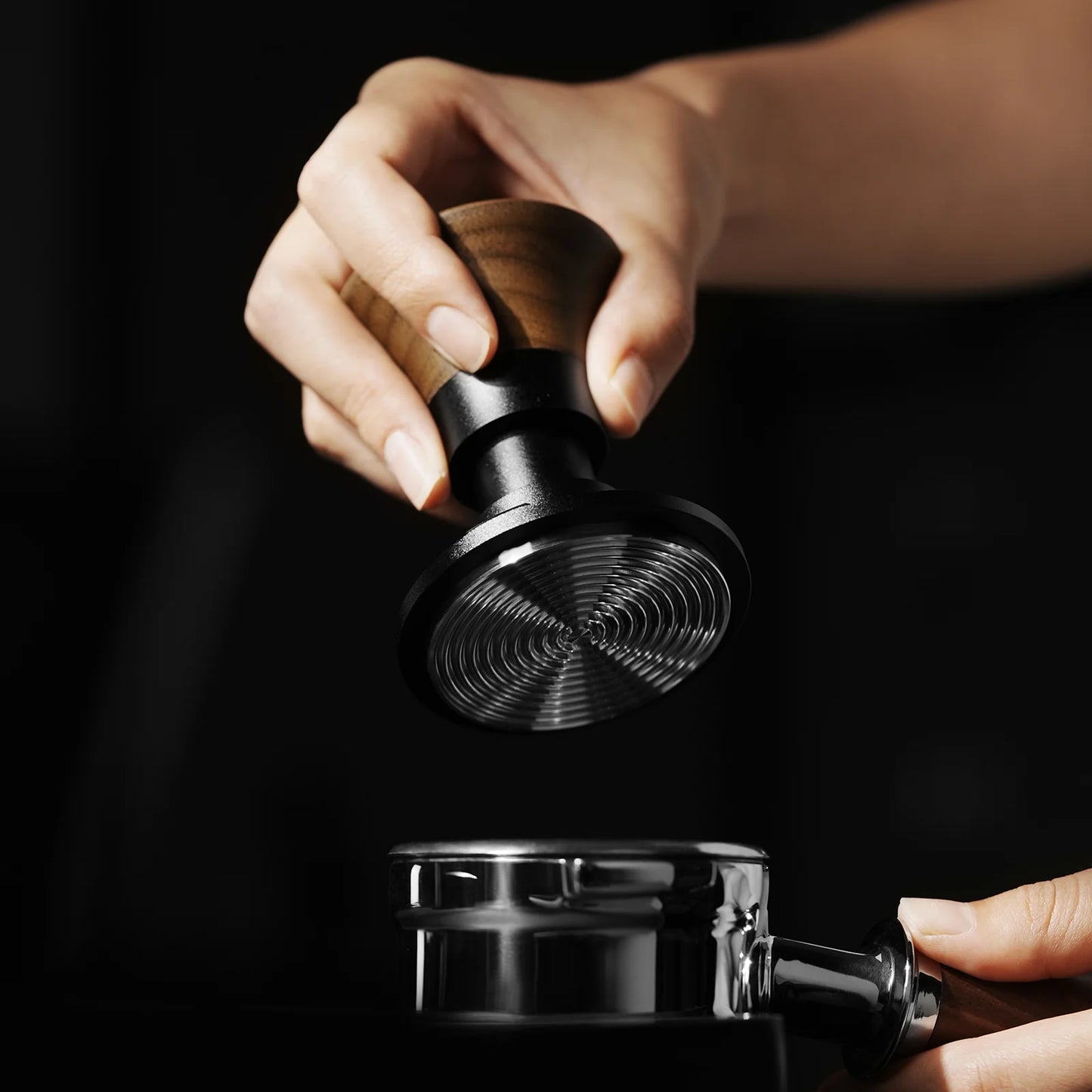 A hand grips the MHW-3BOMBER Armor Series Coffee Tamper 2.0 with a wooden handle, highlighting its consistent pressure and adjustable tamping depth, as it hovers above a metal portafilter against a dark background, emphasizing the polished precision of these MHW-3BOMBER tools.