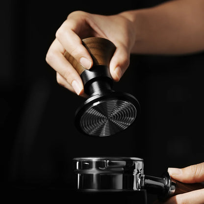 A hand grips the MHW-3BOMBER Armor Series Coffee Tamper 2.0 with a wooden handle, highlighting its consistent pressure and adjustable tamping depth, as it hovers above a metal portafilter against a dark background, emphasizing the polished precision of these MHW-3BOMBER tools.