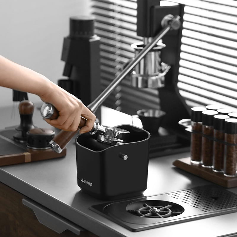On a modern kitchen countertop, someone uses a coffee grinder and a manual espresso machine alongside spice jars and the MHW-3BOMBER Coffee Espresso Square Knock Box. Sunlight filters through blinds, casting shadows across the easy-to-clean, durable workspace.
