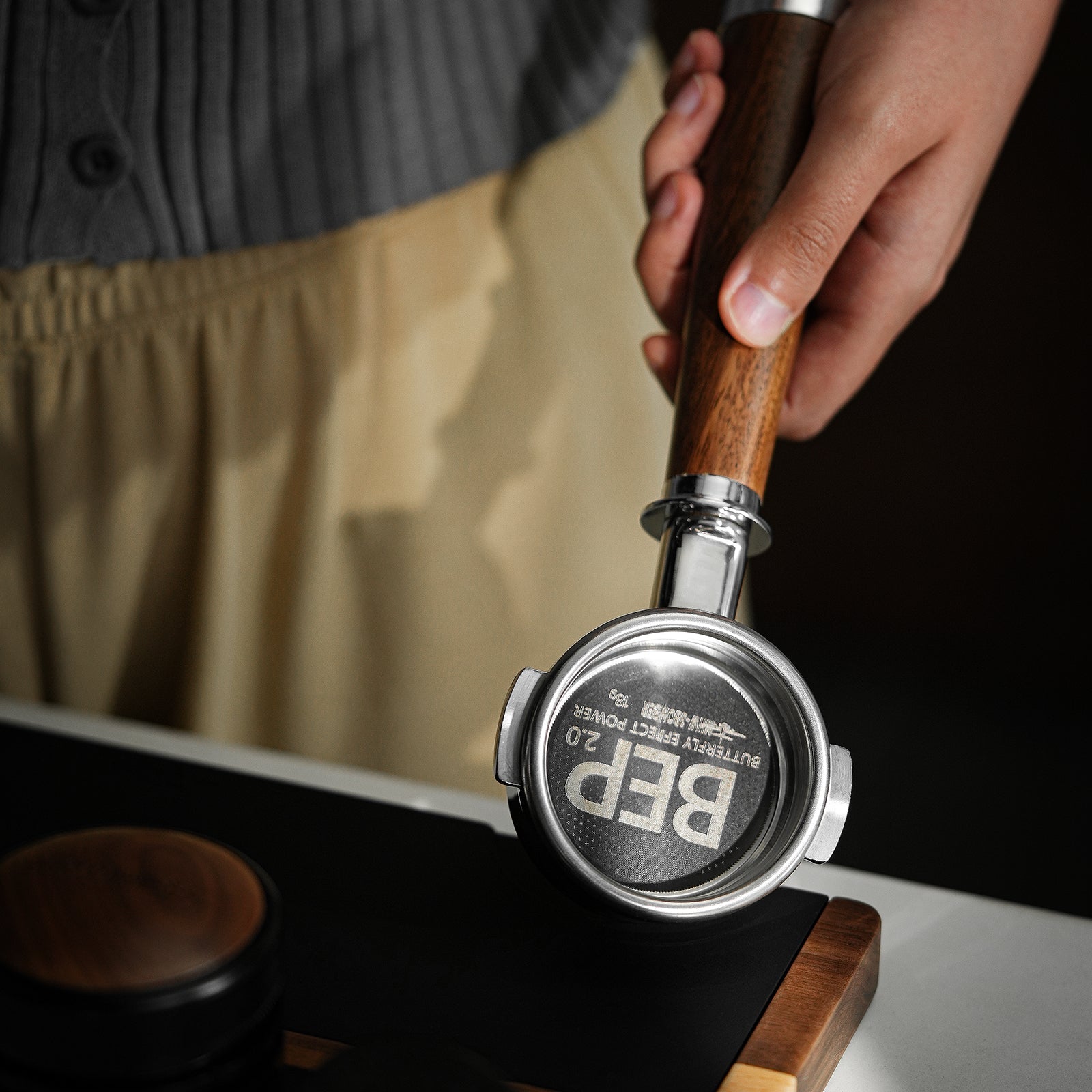 A person in a grey top and beige pants uses the MHW-3BOMBER BEP Butterfly Coffee Filter Basket 2.0 (58.5mm) with dual-layer filtration, holding a wooden-handled portafilter to press ground coffee while a tamper rests on a dark surface nearby.