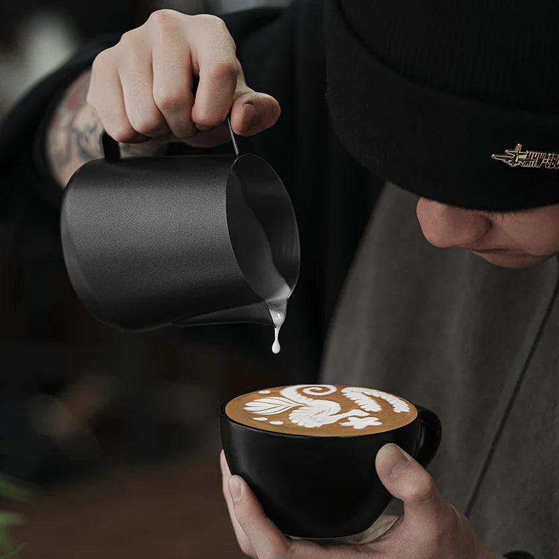 MHW-3BOMBER GT Milk Pitcher in use. Black pitcher pouring milk into black cup with latte art. Barista visible. 