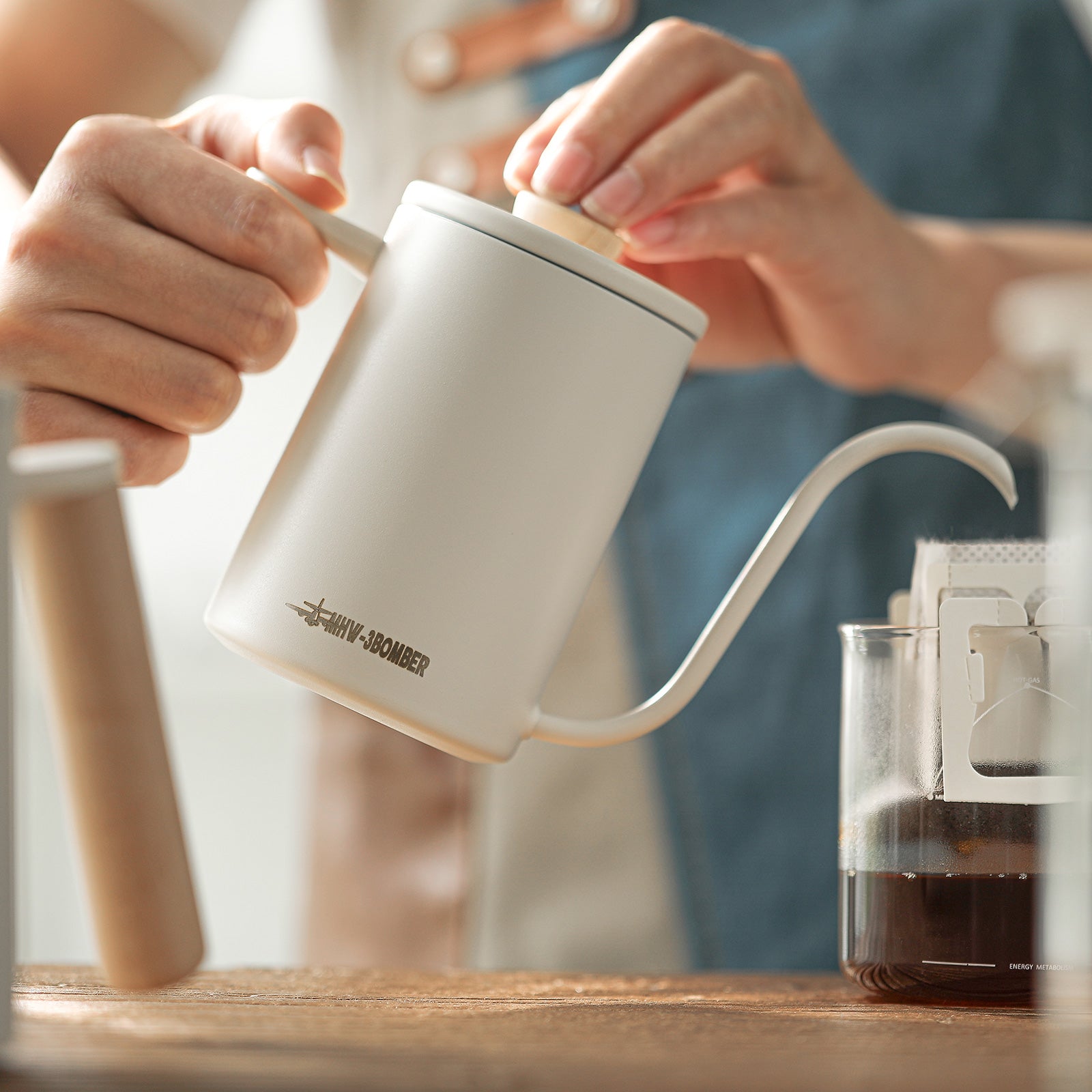 A person gracefully pours hot water from the MHW-3BOMBER Planet Hand Brewing Kettle into a glass, creating a serene morning coffee ritual on a wooden table—each drip adding to the tranquility.
