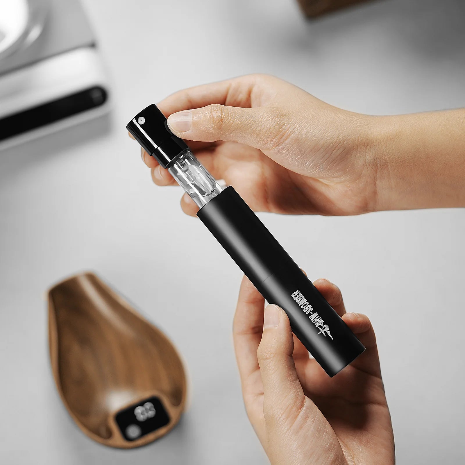 Two hands elegantly hold the MHW-3BOMBER Ovo Coffee Scale & Spray Bottle Set with silver accents over a blurred backdrop, while a wooden dish and espresso scale nearby add brewing sophistication to the scene.