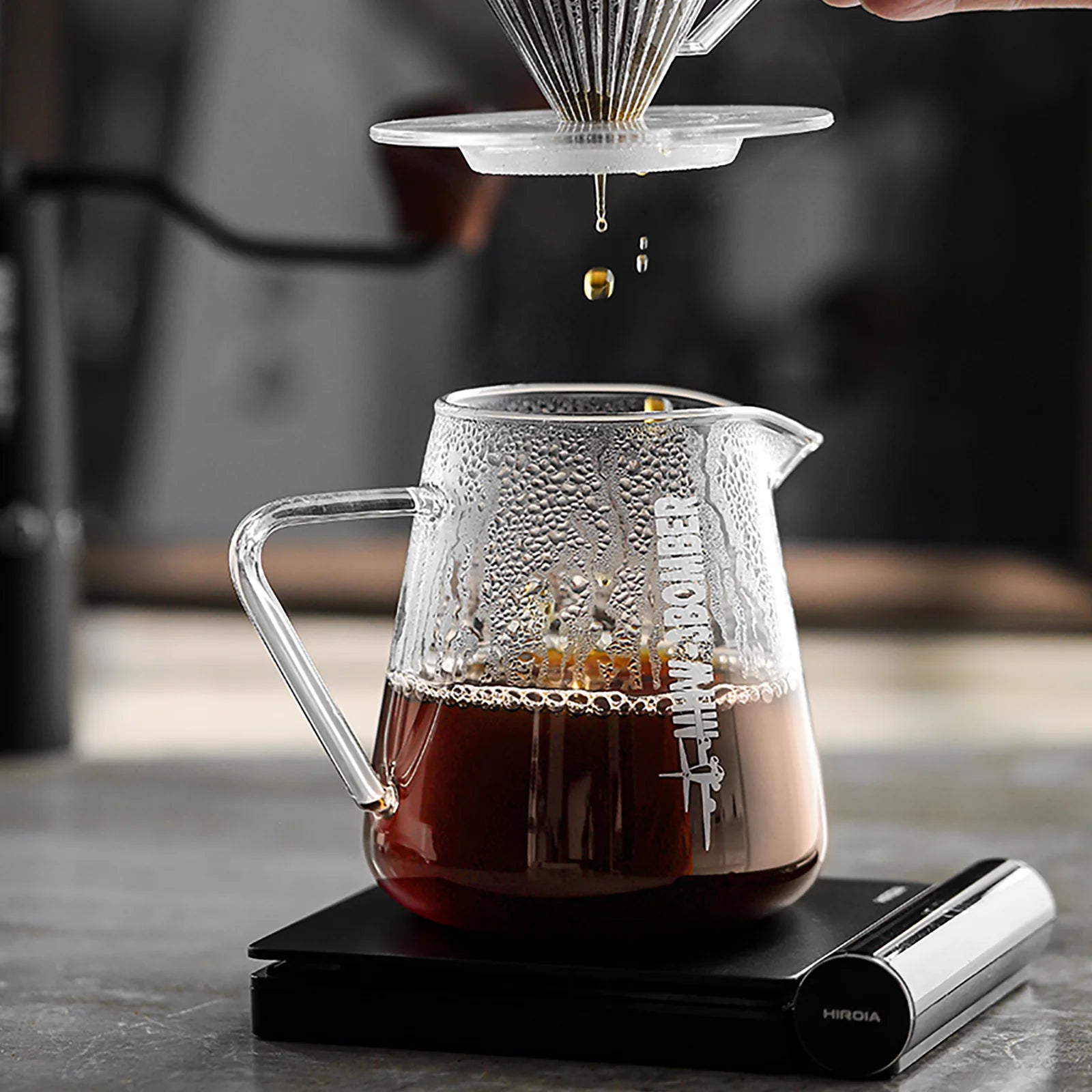 The MHW-3BOMBER Elf Coffee Server, crafted from borosilicate glass, sits on an electronic scale filled with dark coffee. Above it, a hand holds a pour-over dripper, seamlessly dripping coffee into the decanter. Light steam fogs the surface as a grinder rests in the background.