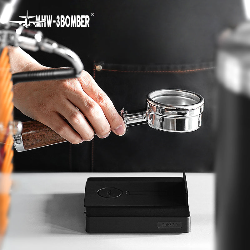 A person in a dark apron holds a wooden-handled coffee portafilter with a transparent bottom near an espresso machine, while the MHW-3BOMBER Cube Coffee Scale 2.0 Mini rests on the counter, ensuring each brew is impeccable.
