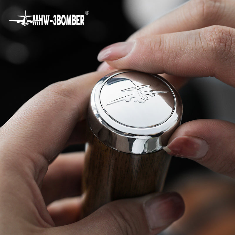 Close-up of hands holding a MHW-3BOMBER gear shifter crafted from natural walnut wood, featuring an engraved airplane logo on top. This unique piece, labeled IW-3BOMBER, is enhanced with sleek stainless steel accents.