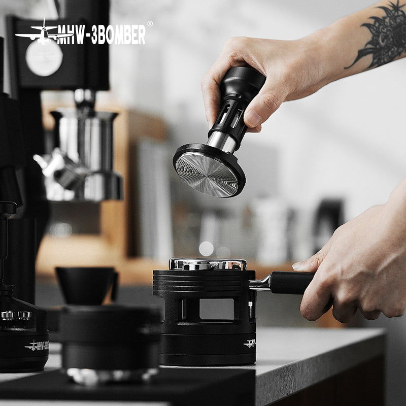 MHW-3BOMBER 58mm espresso tamper in use. Black metal, silver tamping base. Tamping coffee in portafilter on counter.