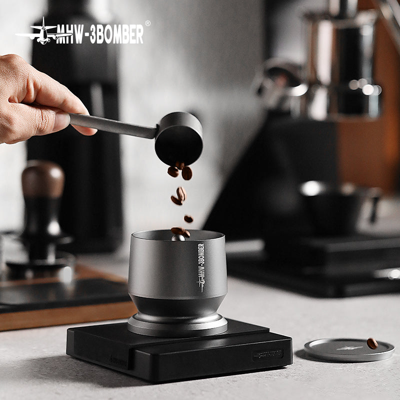 A hand expertly pours coffee beans into a sleek metal container on a digital scale, highlighting brewing precision. The MHW-3BOMBER Espresso Coffee Powder Dosing Funnel and other tools blur in the background, epitomizing the art of baristas at work.