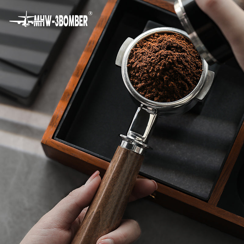 A hand holds an MHW-3BOMBER Bottomless Portafilter filled with fresh coffee grounds above a wooden box made from natural walnut. Another hand reaches to adjust the coffee, with the MHW-3BOMBER logo visible in the top left corner.