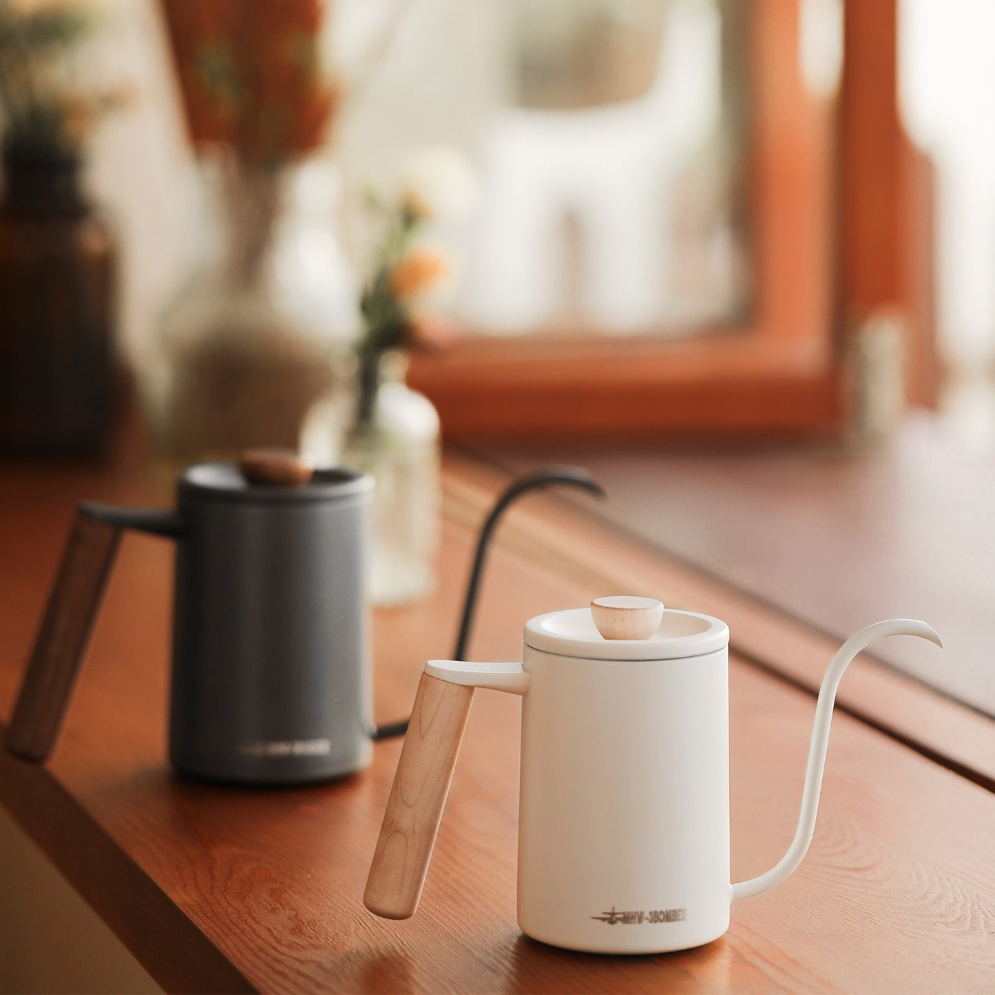 A white MHW-3BOMBER Planet Hand Brewing Kettle with a wooden handle sits on a wooden surface, ideal for drip coffee. A similar black kettle is blurred in the background near a window, with out-of-focus plants visible outside.