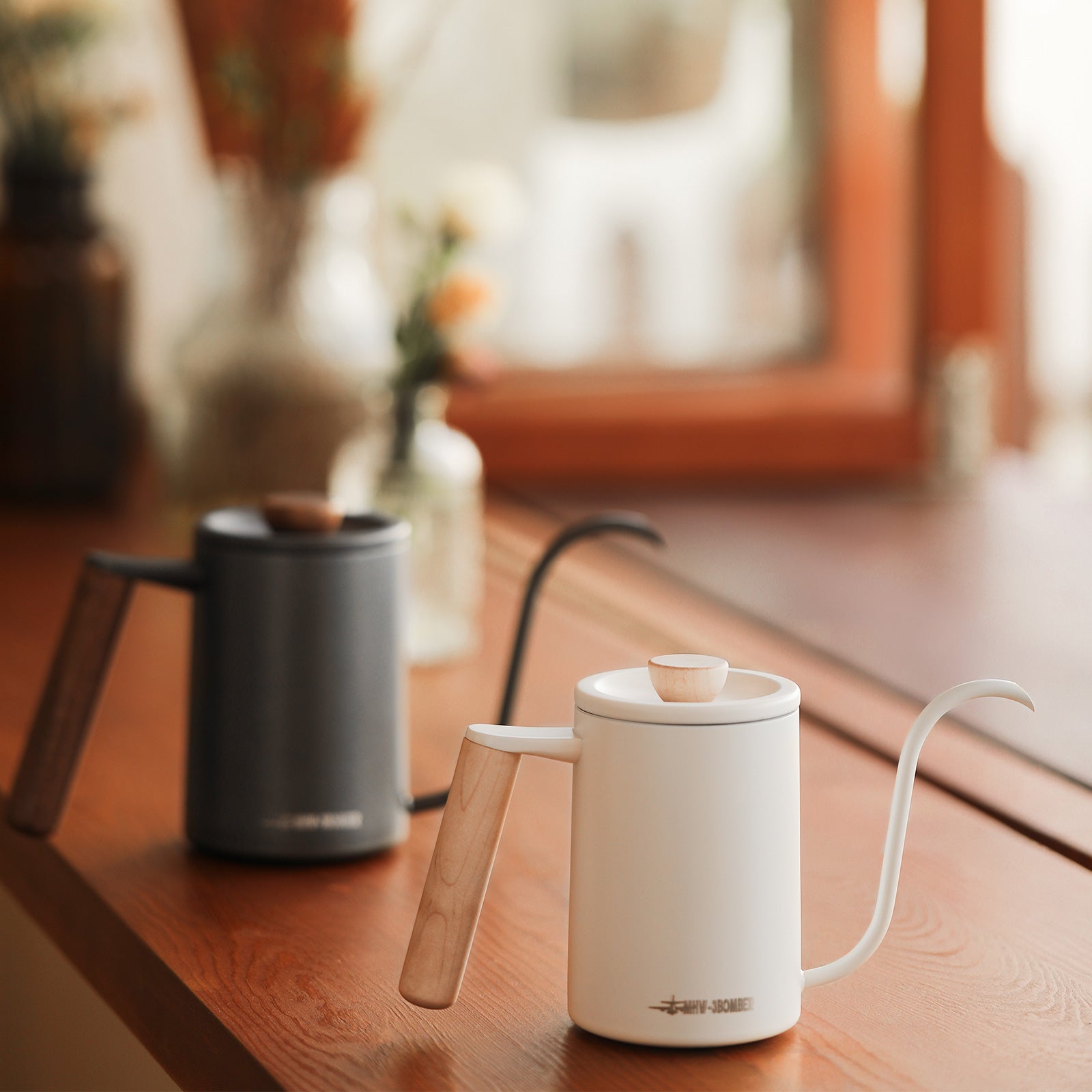 A white MHW-3BOMBER Planet Hand Brewing Kettle with a wooden handle sits on a wooden surface, ideal for drip coffee. A similar black kettle is blurred in the background near a window, with out-of-focus plants visible outside.