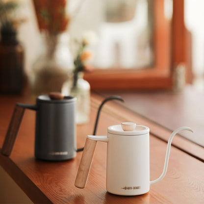 A white MHW-3BOMBER Planet Hand Brewing Kettle with a wooden handle sits on a wooden surface, ideal for drip coffee. A similar black kettle is blurred in the background near a window, with out-of-focus plants visible outside.