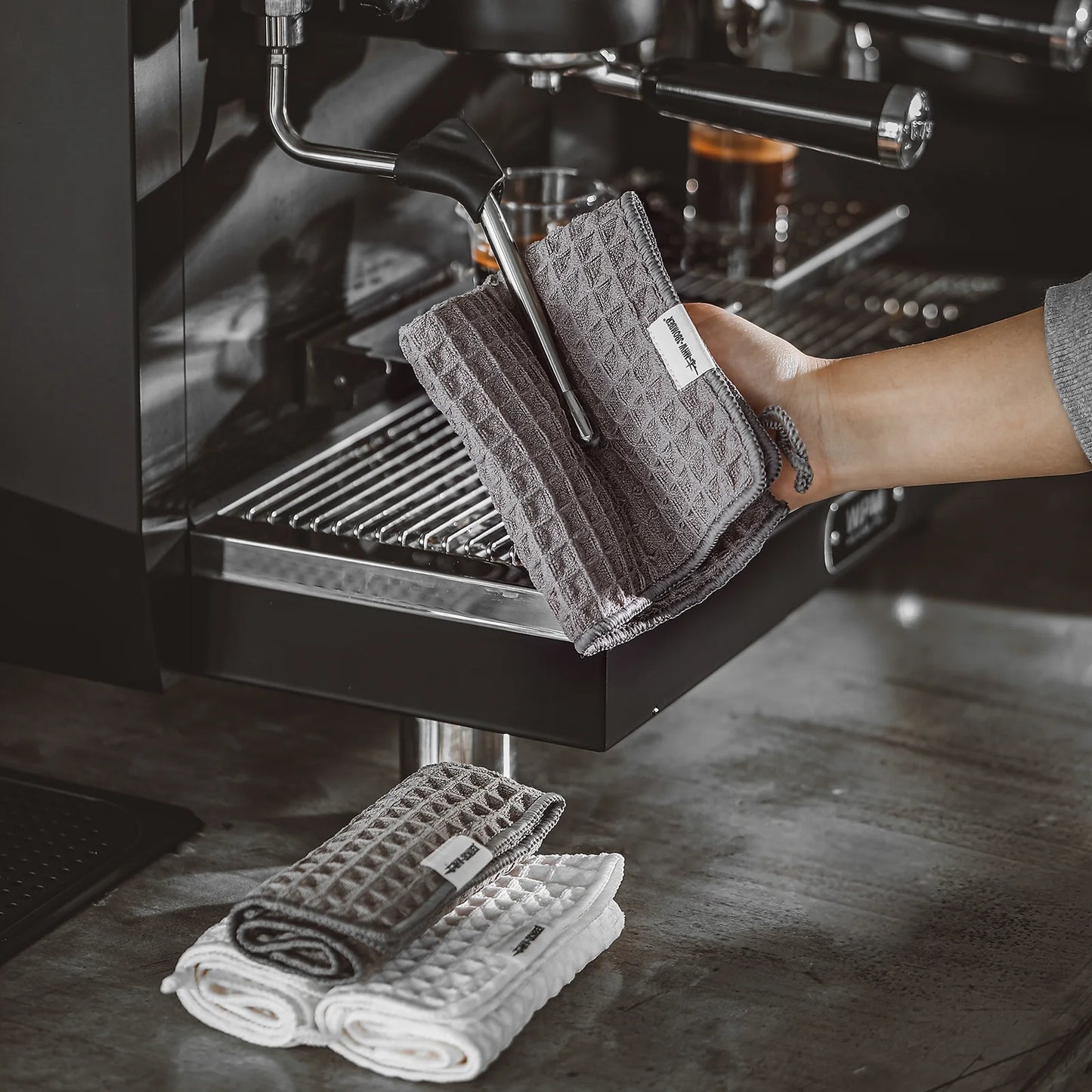 MHW-3BOMBER gray waffle towel used on espresso machine, plus stacked towels. Absorbent, quick-drying microfiber for baristas.