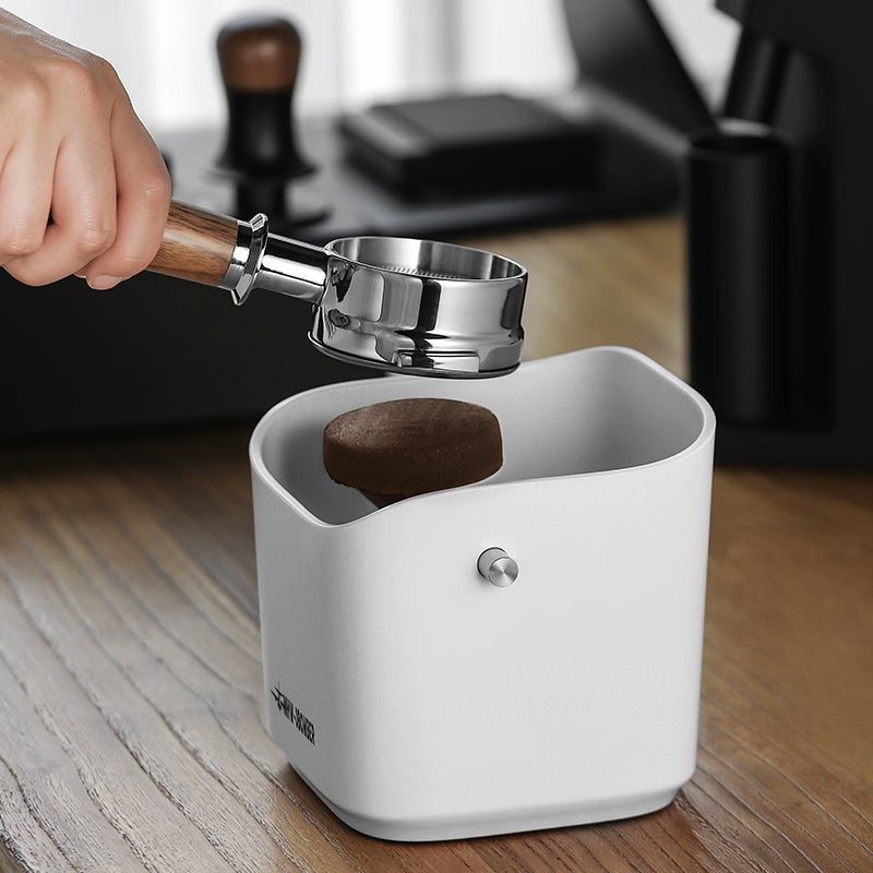 A hand grips a portafilter with a used espresso puck ready to drop into the MHW-3BOMBER Coffee Espresso Square Knock Box on a wooden countertop, with a blurred espresso machine in the background.