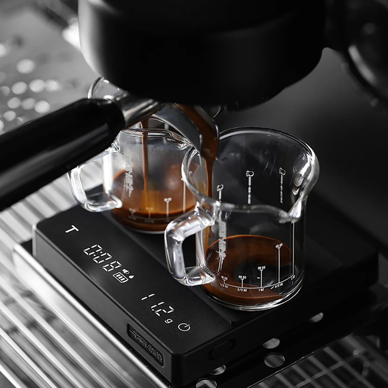 Close-up of an MHW-3BOMBER Espresso Scale Cube Coffee Scale 2.0 with an espresso machine pouring coffee into measuring cups. The scale, USB-rechargeable with a sleek metallic grid surface, displays weight or timing measurements on its screen.