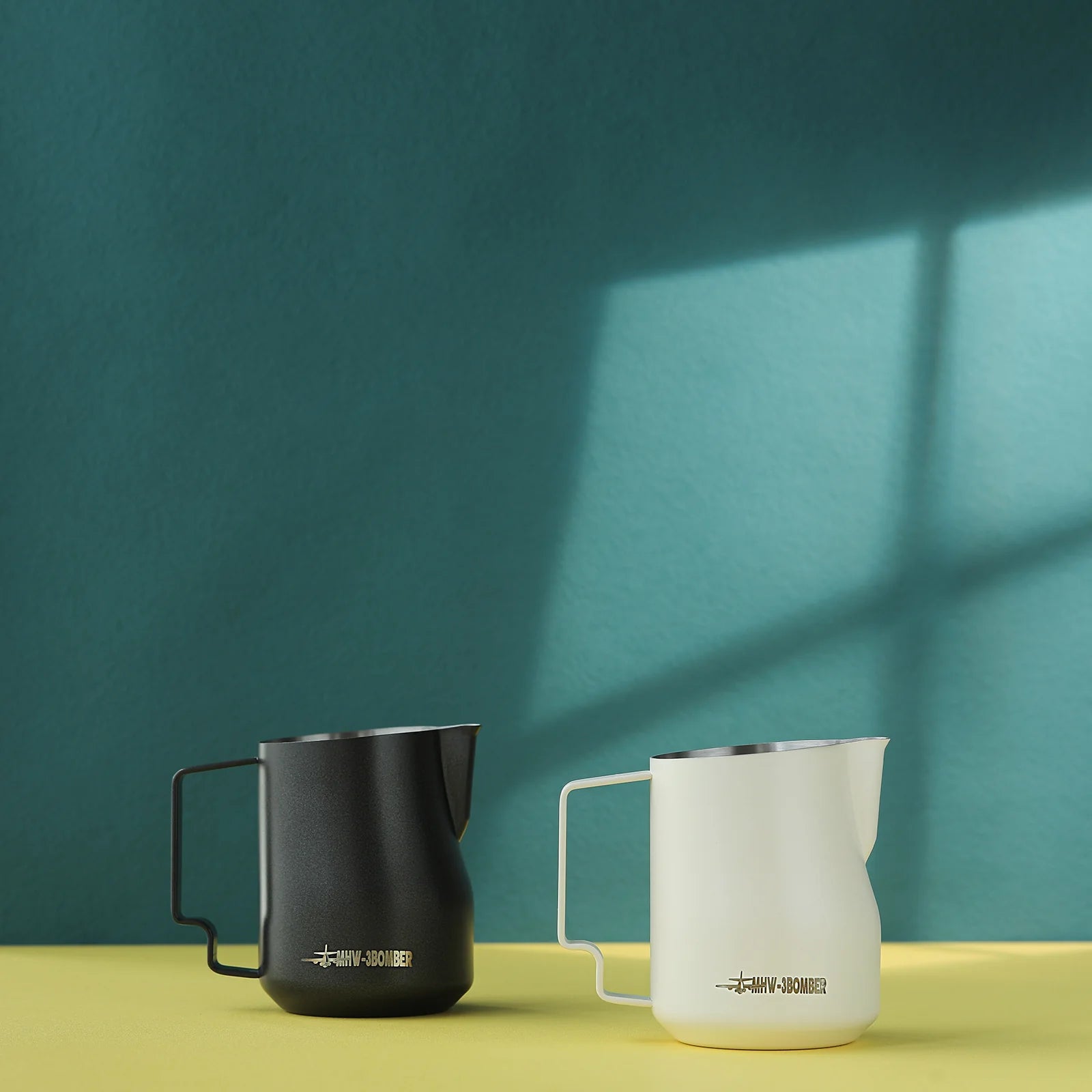 Two MHW-3BOMBER Turbo Milk Pitchers, crafted from stainless steel with a matte finish in black and white, are displayed on a yellow surface against a teal background. Sunlight casts shadows, highlighting their modern design and precision for perfect latte art.