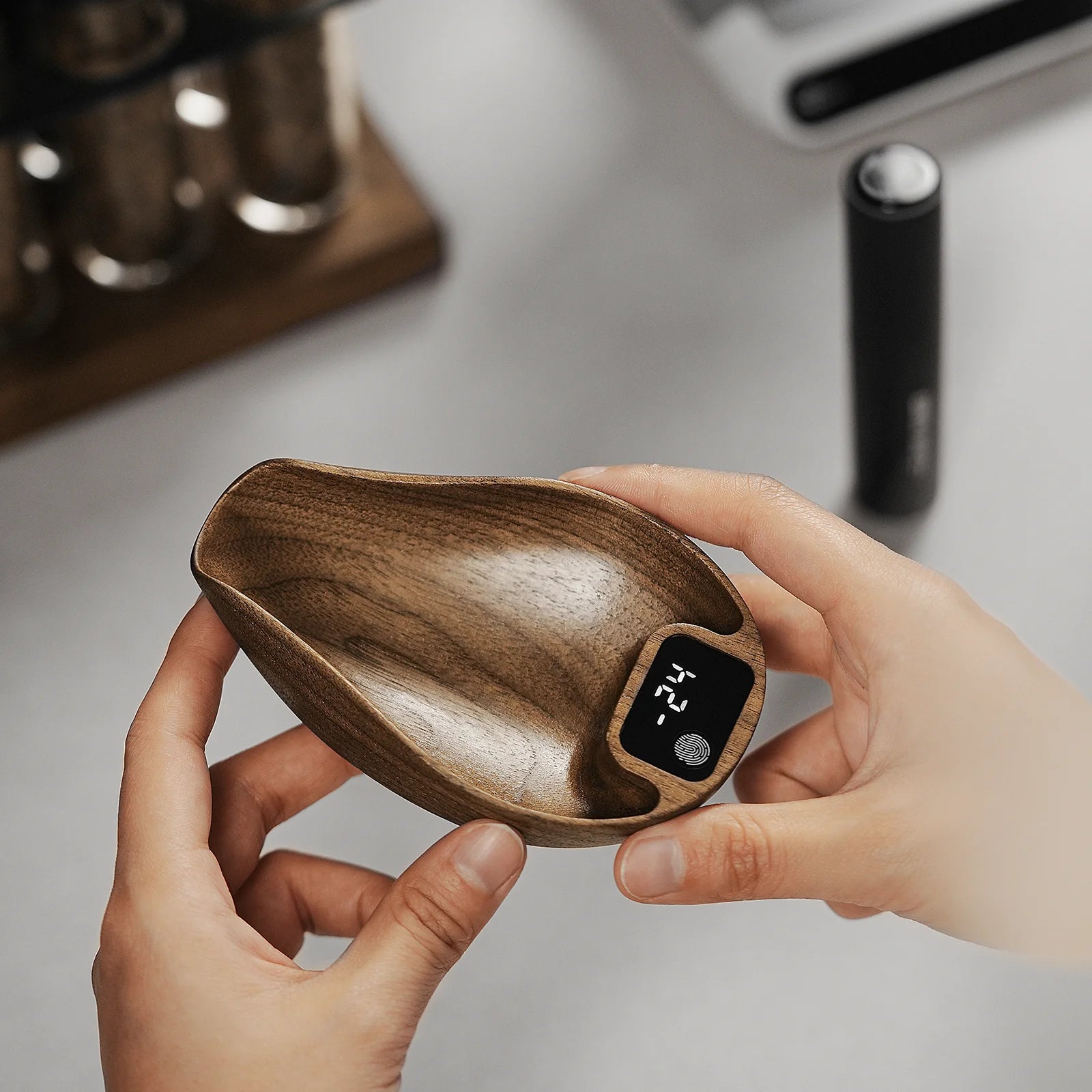 A person holds a small, curved wooden bowl with a black digital thermometer inside, highlighting precision in brewing. Blurred background containers and a cylindrical object evoke tools such as the MHW-3BOMBER Ovo Coffee Scale & Spray Bottle Set.