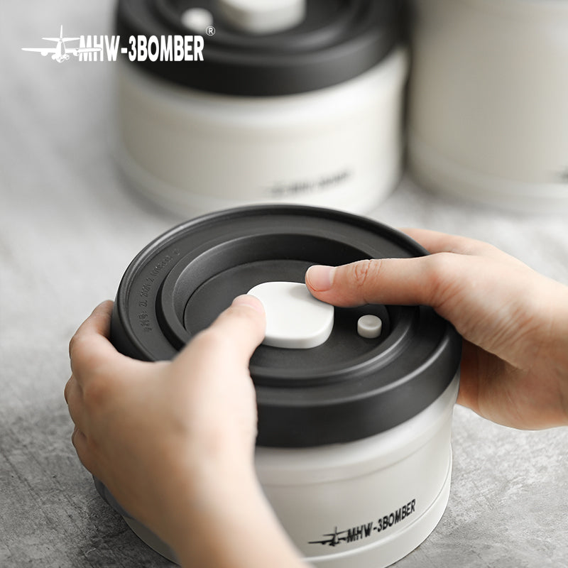 MHW-3BOMBER vacuum canister, white and gray. Hands pressing the air pump on the lid. Coffee storage container.