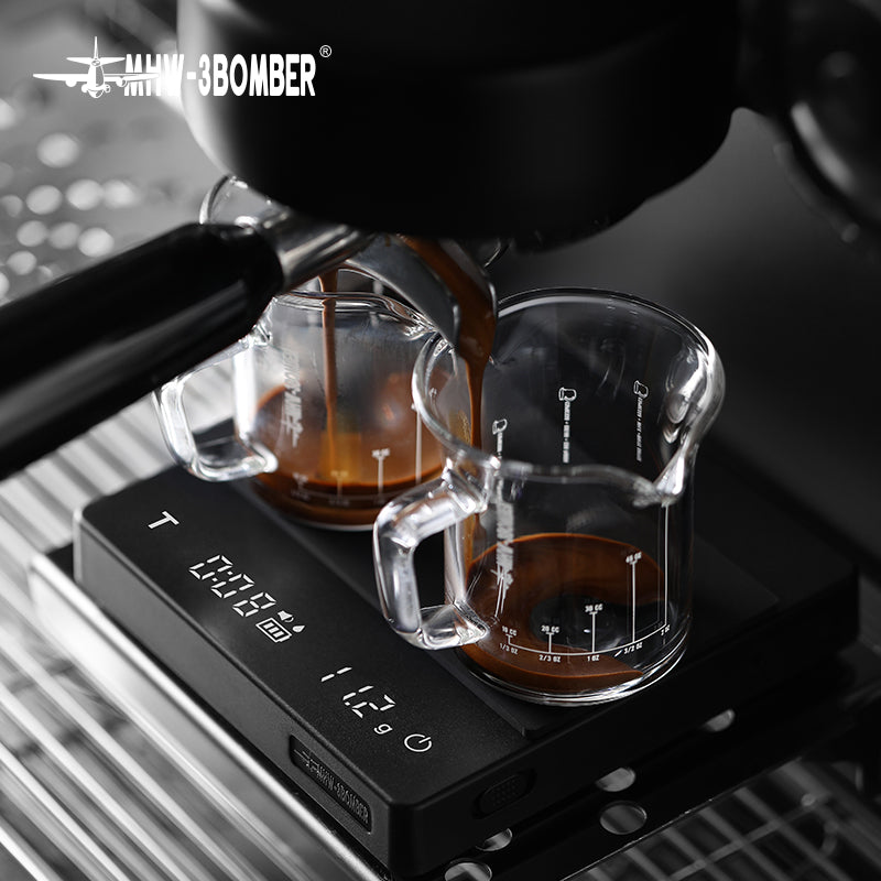 Close-up of espresso flowing into two small glass pitchers on a MHW-3BOMBER Cube Coffee Scale 2.0 Mini. The display shows numbers as the portafilter hovers above, ensuring every drop counts with USB rechargeable convenience.