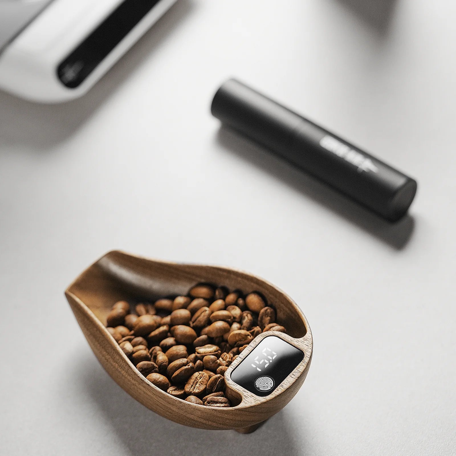 An MHW-3BOMBER Ovo Coffee Scale & Spray Bottle Set, featuring a wooden bowl of coffee beans and a digital timer, graces a light surface. Part of the cylindrical spray bottle and the espresso scale are visible in the background, encapsulating the brewing essence.