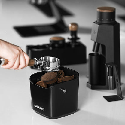 A hand holds a portafilter filled with coffee grounds above the MHW-3BOMBER Coffee Espresso Square Knock Box. In the background, various coffee tools like a grinder and tamper rest on the white counter, ready for the next brew.
