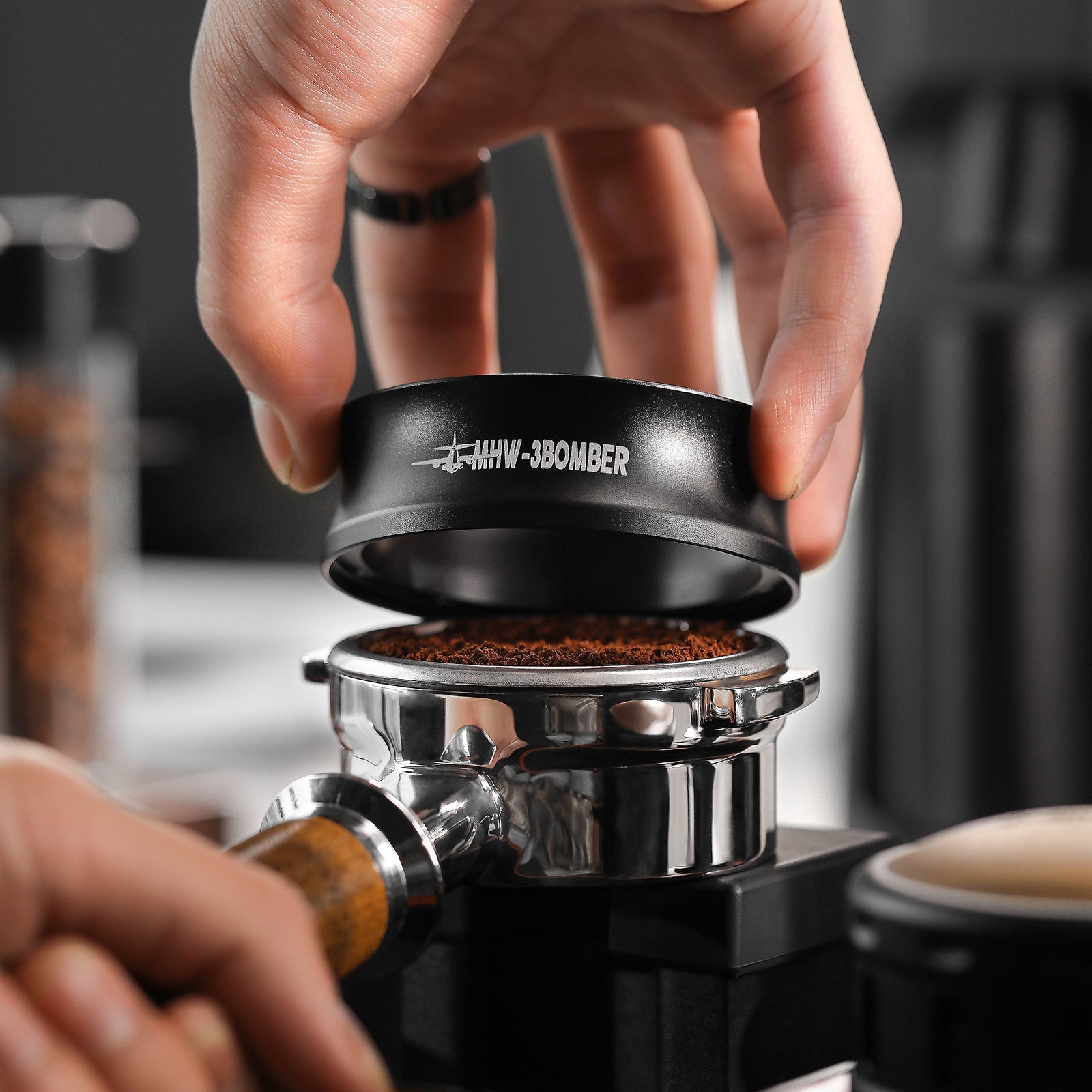 A hand skillfully presses coffee grounds in a portafilter with a black tamper, using the MHW-3BOMBER Espresso Magnetic Coffee Dosing Funnel for precise dosing. The scene exudes an ambiance of meticulous coffee-making and dedication to quality.
