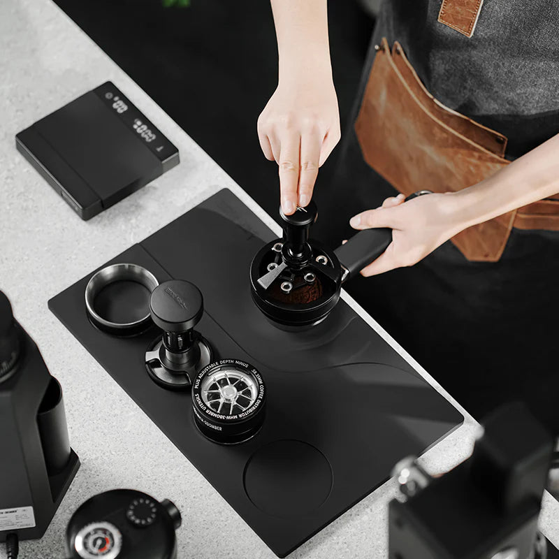 MHW-3BOMBER black silicone tamping base with portafilter, tamper, and distributor slots. Person tamping.