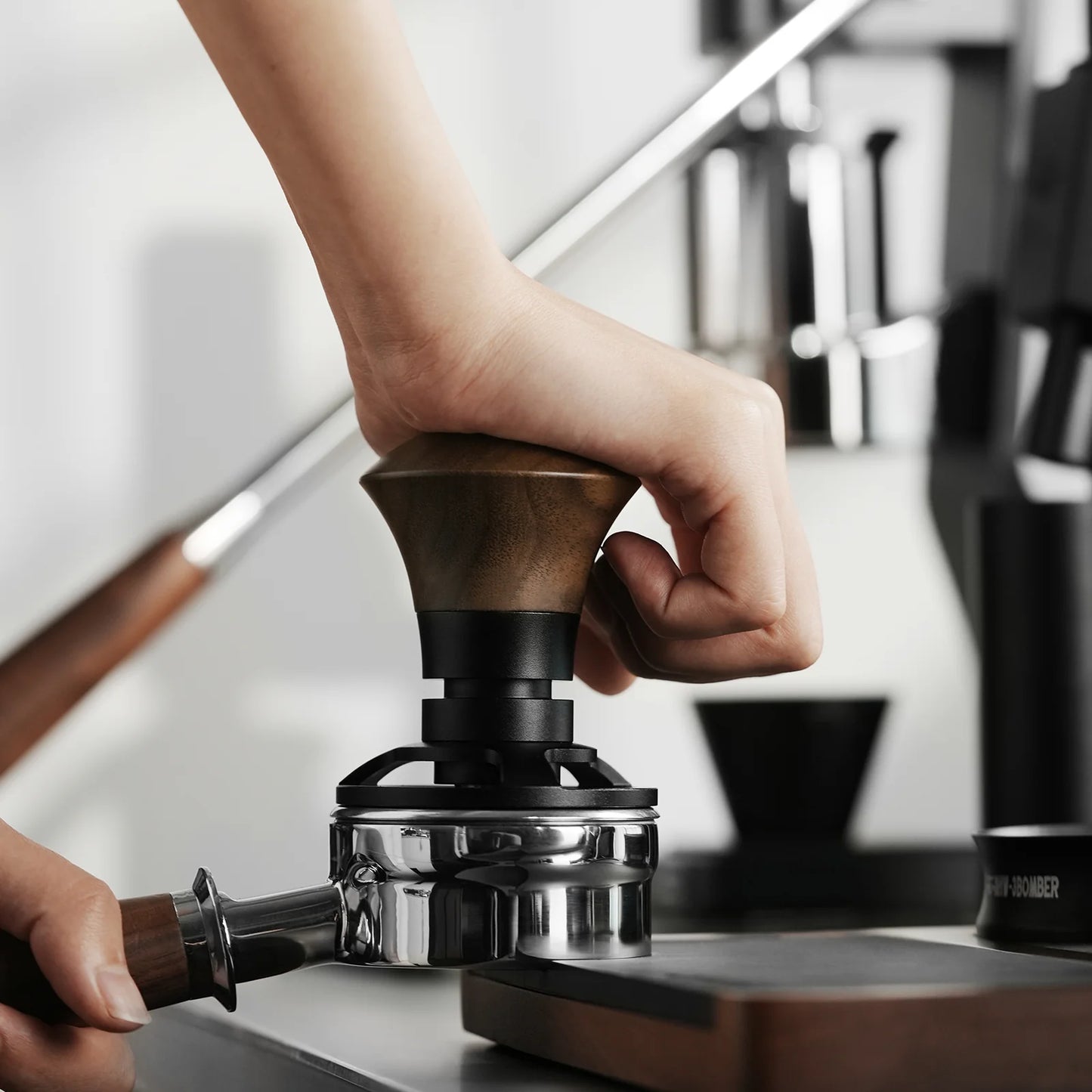 A hand expertly uses the MHW-3BOMBER Armor Series Coffee Tamper 2.0 with its elegant wooden handle to press coffee grounds in a portafilter, capturing the magic of morning rituals against a backdrop of artfully blurred kitchen equipment.