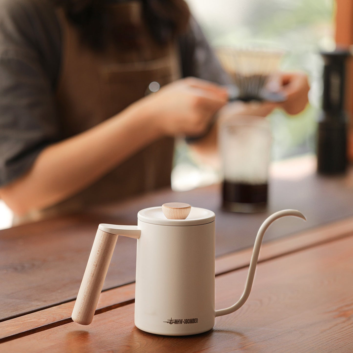 The MHW-3BOMBER Planet Hand Brewing Kettle, featuring a wooden handle and lid, sits elegantly on a table. In the blurred background, someone prepares pour-over coffee using a drip kettle, coffee dripper, and glass carafe.