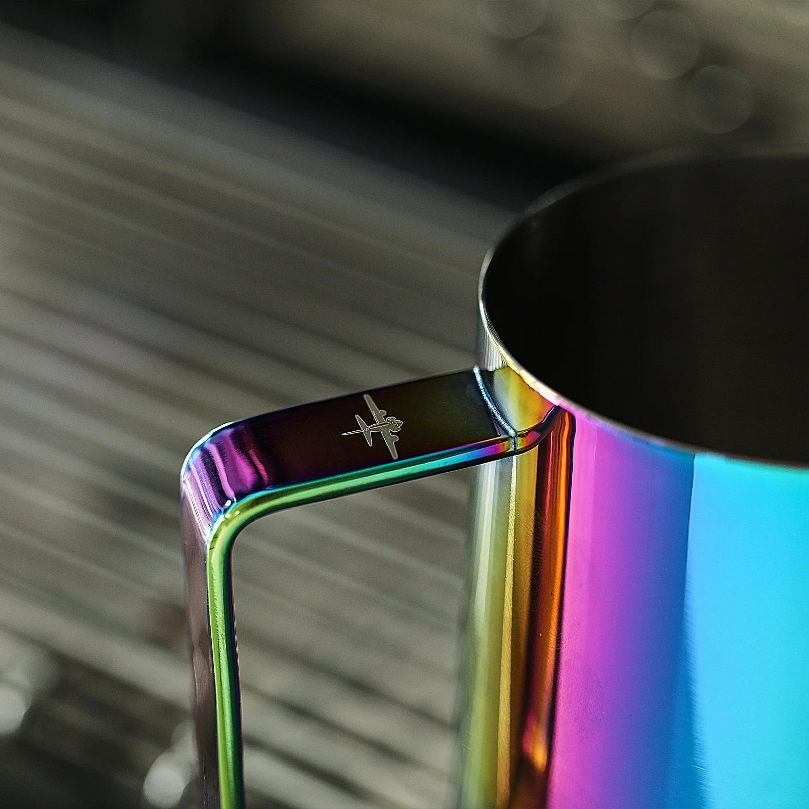 The MHW-3BOMBER Latte Art Pitcher 5.0 Colorful is a shiny, iridescent stainless steel pitcher with a sleek handle, crocodile spout, and an airplane logo. Its vibrant metallic surface reflects various hues against a blurred background.