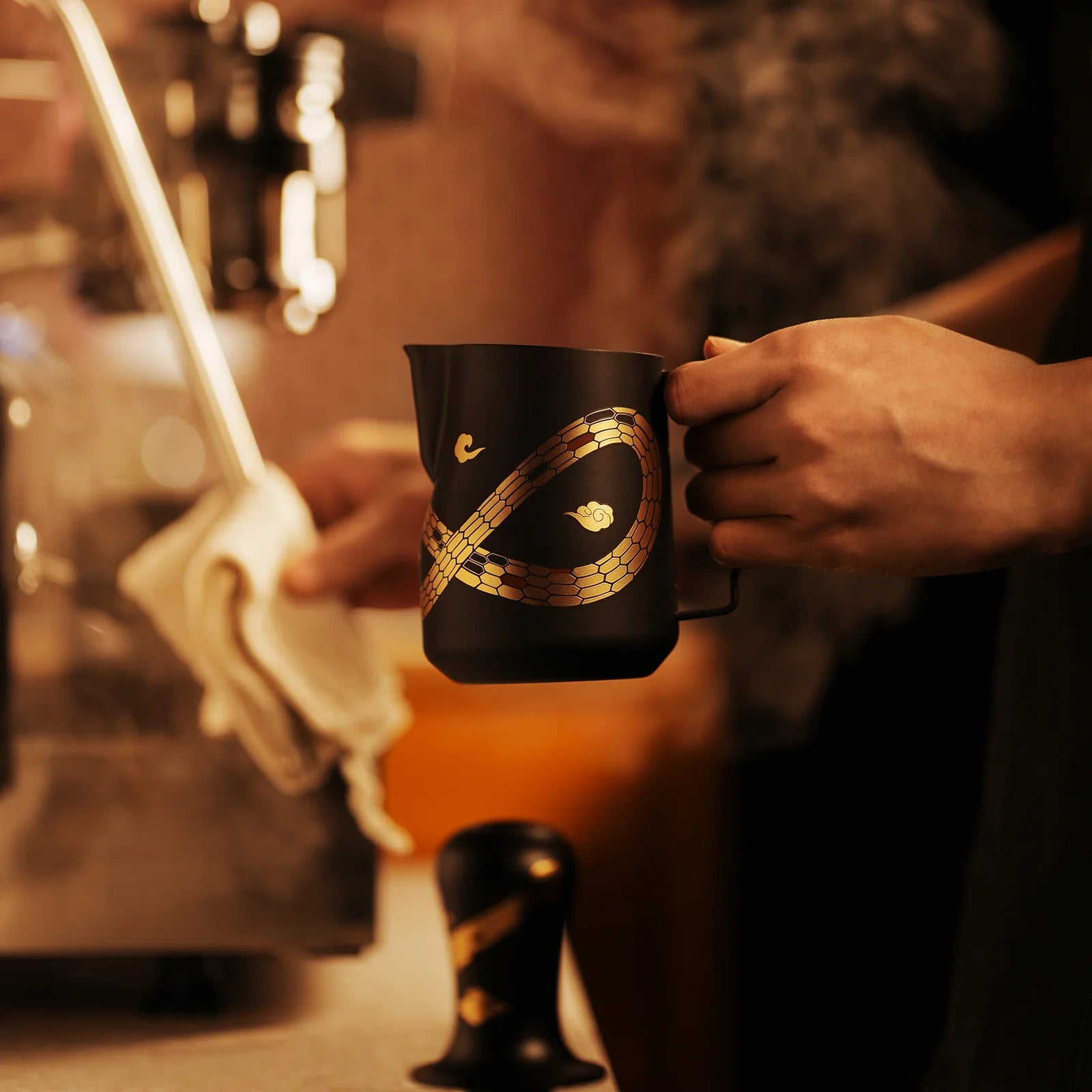 A person holds an MHW-3BOMBER Turbo Milk Pitcher-Year of the Snake Limited Edition, black with gold leaf patterns. Nearby, a coffee tamper and espresso machine release steam against a softly lit, blurred background.