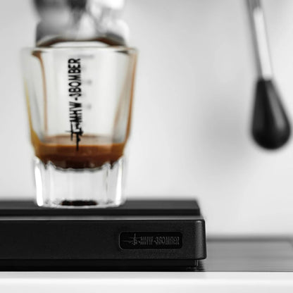 Close-up of an espresso being pulled from a machine, flowing into a glass on the MHW-3BOMBER Espresso Scale Cube Coffee Scale 2.0, featuring high precision and USB rechargeability. The background is out of focus, emphasizing the brewing process.