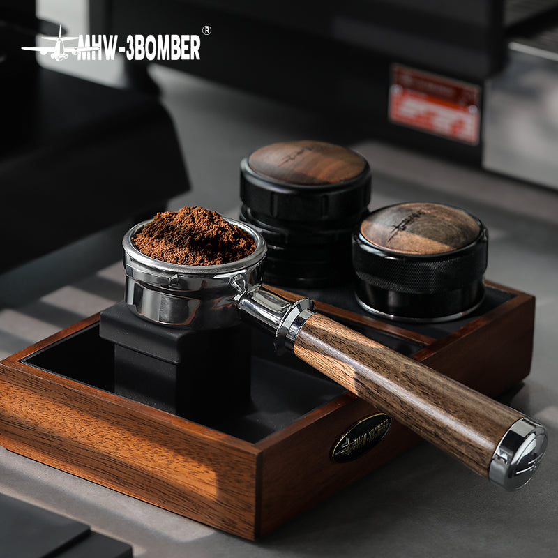 An MHW-3BOMBER Bottomless Portafilter filled with ground coffee rests on a polished wooden stand. Nearby, two sleek, black and natural walnut wood tampers are neatly arranged on a gray surface, exuding elegance and precision.