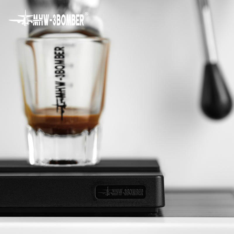 Close-up of an espresso shot pouring into a MHW-3BOMBER glass resting on a precise, USB rechargeable MHW-3BOMBER Cube Coffee Scale 2.0 Mini. Part of an espresso machines portafilter is visible on the right.