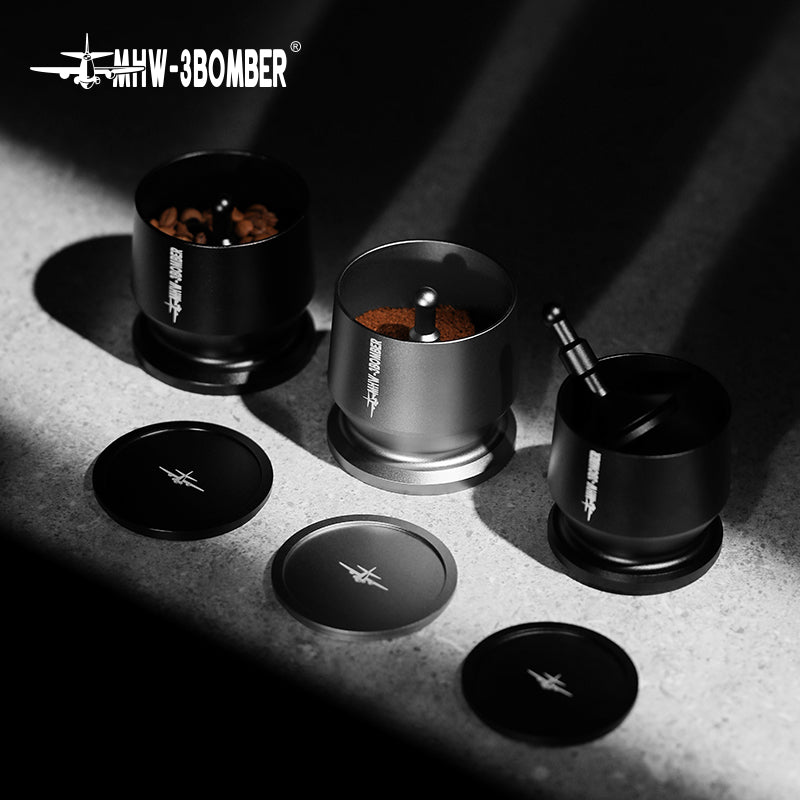Three black metal MHW-3BOMBER grinders are on a stone surface at various coffee grinding stages: whole beans, partially ground, and fully ground. Lids with MHW-3BOMBER logos are in front, ideal for baristas using the Espresso Coffee Powder Dosing Funnel.