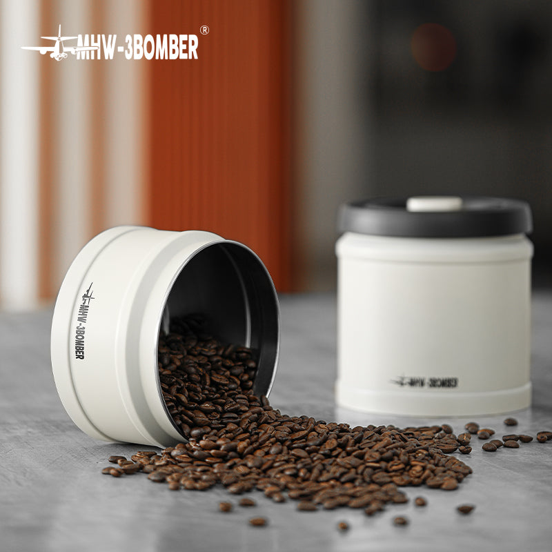 MHW-3BOMBER white vacuum coffee canister with black lid, beans spilling. Stylish storage for fresh coffee.