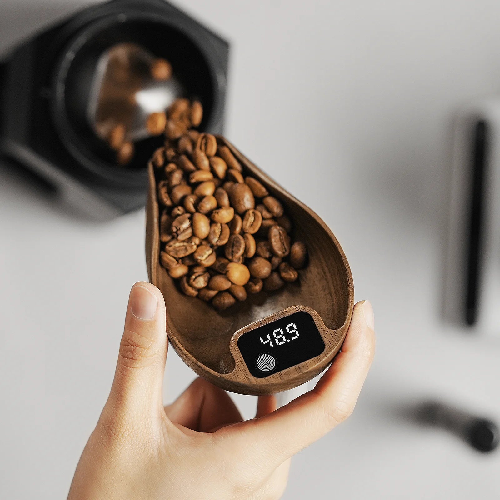 A hand holds a wooden coffee scoop filled with beans on an MHW-3BOMBER Ovo Coffee Scale & Spray Bottle Set, displaying 48.9. In the blurred background, a coffee grinder is on the marble counter, ready to enhance your brewing process.