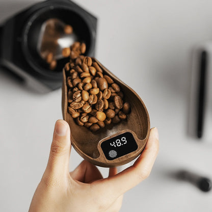 A hand holds a wooden coffee scoop filled with beans on an MHW-3BOMBER Ovo Coffee Scale & Spray Bottle Set, displaying 48.9. In the blurred background, a coffee grinder is on the marble counter, ready to enhance your brewing process.