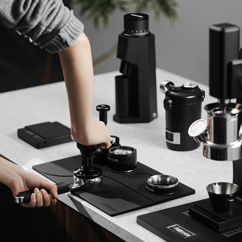 MHW-3BOMBER black tamping base with portafilter and tamper. Organizes coffee accessories on a kitchen counter. 