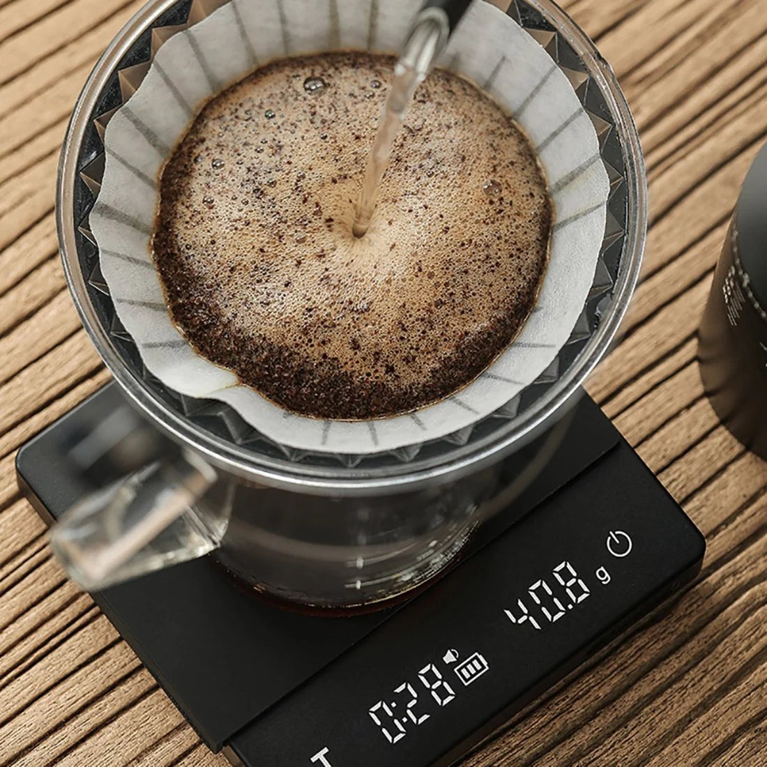 A pour-over coffee setup showcases hot water flowing over grounds in a paper filter within a glass dripper on an MHW-3BOMBER Espresso Scale Cube Coffee Scale 2.0, showing 40.8 grams. The rustic wooden surface beneath complements the aesthetic.